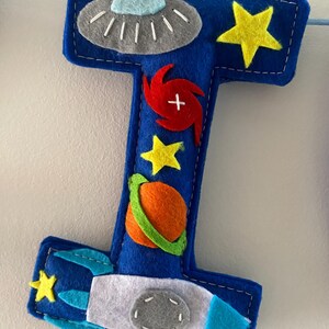 May include: A blue felt letter I decoration featuring space-themed appliqu&eacute;s including a grey UFO, yellow stars, a red spiral galaxy, an orange planet with a green ring, and a white and blue rocket ship, all stitched onto the surface.