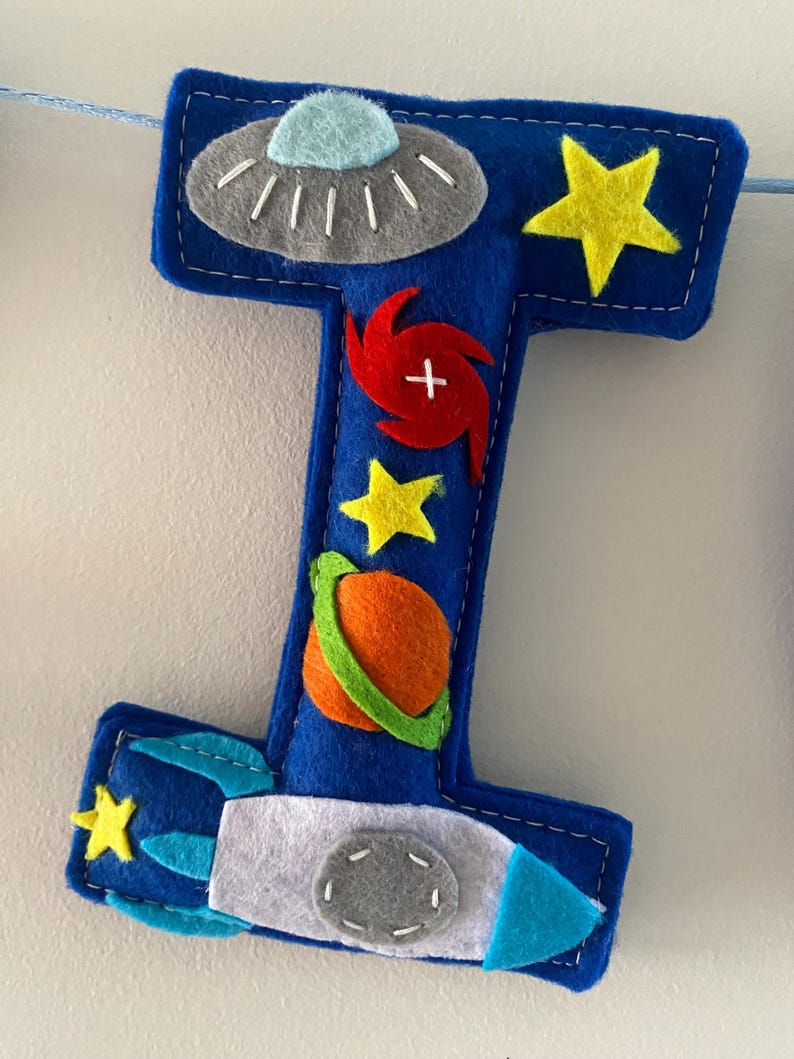 May include: A blue felt letter I decoration featuring space-themed appliqu&eacute;s including a grey UFO, yellow stars, a red spiral galaxy, an orange planet with a green ring, and a white and blue rocket ship, all stitched onto the surface.