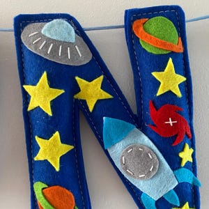 May include: A blue felt letter N decoration featuring space-themed appliqu&eacute;s including a gray UFO, a green planet with an orange ring, yellow stars, a red spiral galaxy, and a light blue rocket ship with a gray circular window, all stitched together.