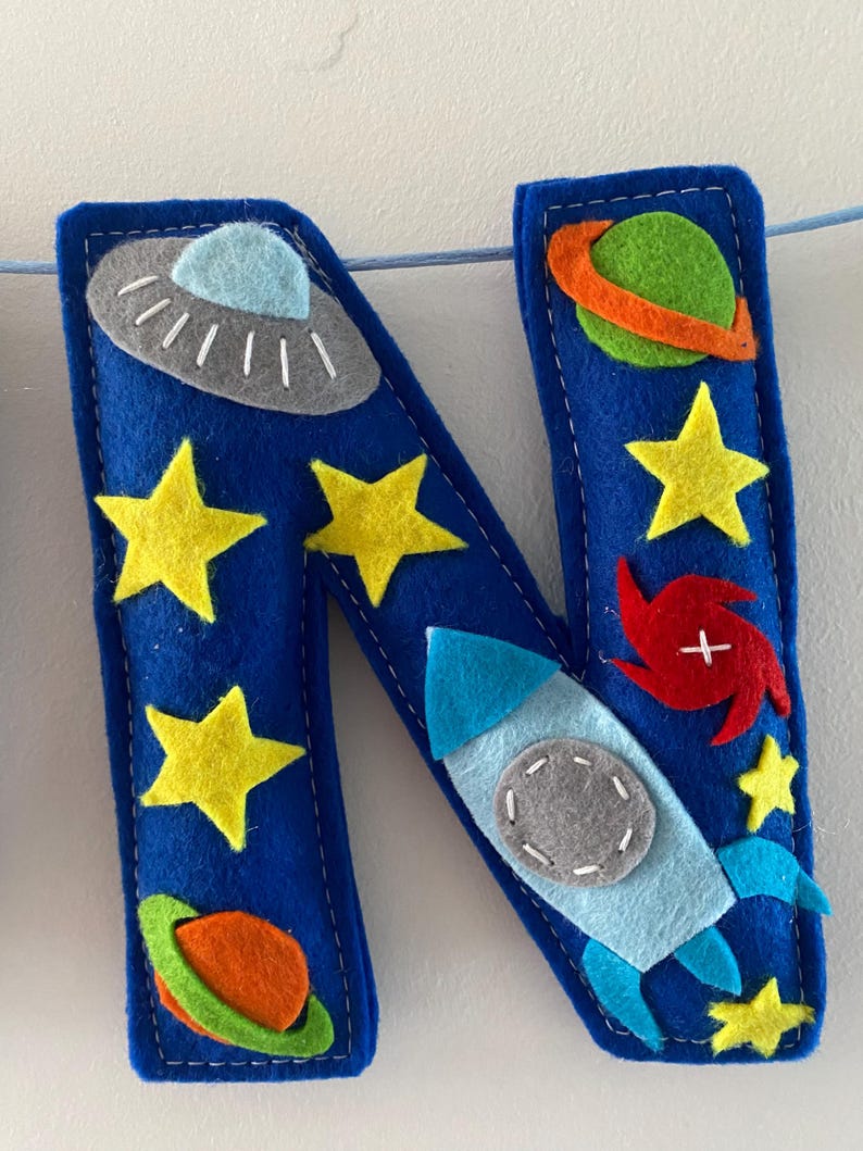 May include: A blue felt letter N decoration featuring space-themed appliqu&eacute;s including a gray UFO, a green planet with an orange ring, yellow stars, a red spiral galaxy, and a light blue rocket ship with a gray circular window, all stitched together.