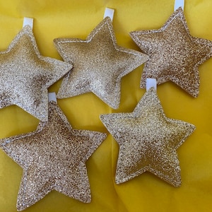 May include: Five gold glitter star-shaped decorations with white stitching. The stars are hanging from white strings.