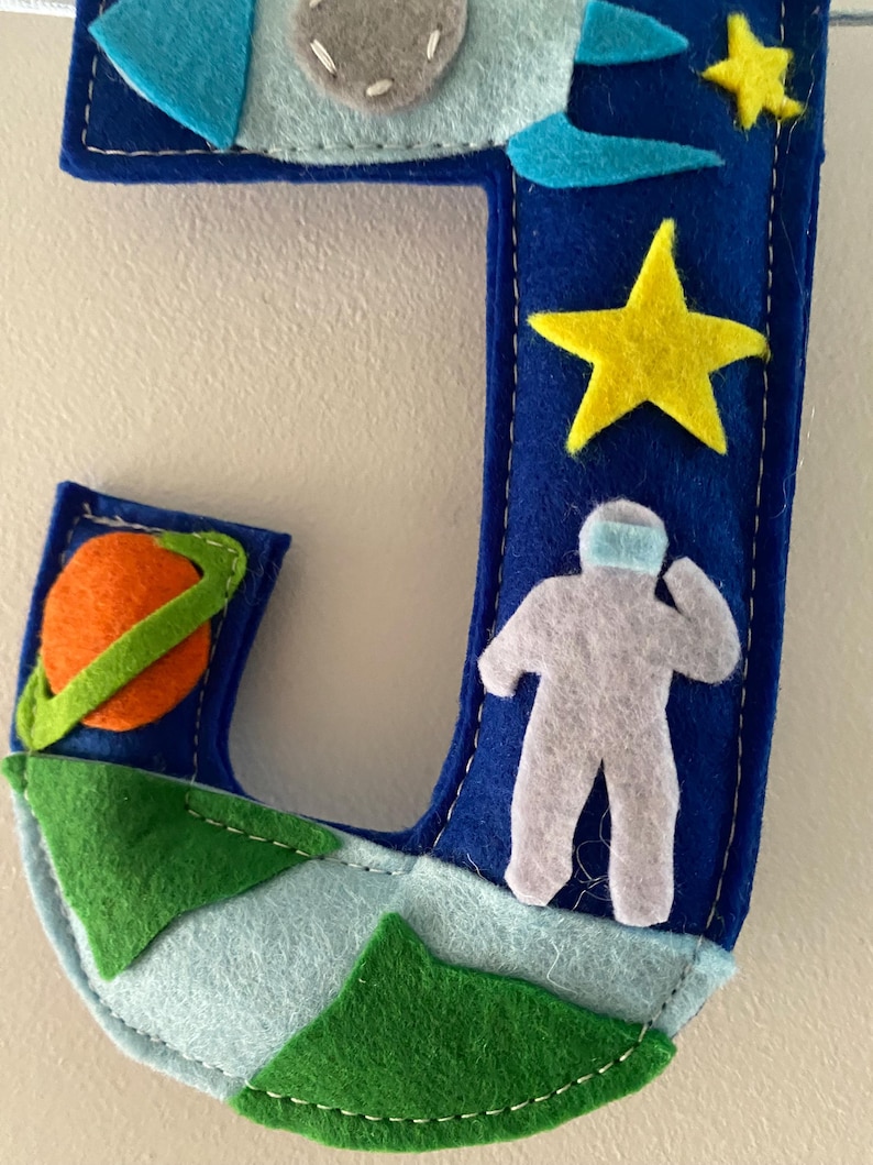May include: A dark blue felt letter J decorated with space-themed appliqu&eacute;s including a light blue rocket, a yellow star, a gray astronaut, an orange planet with a green ring, and green mountain shapes along the bottom edge with white stitching.