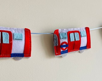 May include: A felt garland featuring three red and white London Underground train cars. Each car has a blue circle with a red circle inside, representing the London Underground logo. The cars are connected by a white string.
