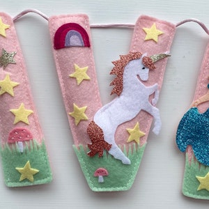 K&ouml;nnte beinhalten: Ein Filzbanner, das den Namen AVA in hellrosa Buchstaben buchstabiert. Das erste A zeigt eine Fee mit Zauberstab, das V ein wei&szlig;es Einhorn mit glitzernder M&auml;hne und Regenbogen, und das zweite A eine Prinzessin in einem blauen Glitzerkleid.