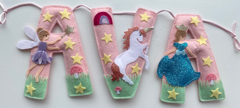 K&ouml;nnte beinhalten: Ein Filzbanner, das den Namen AVA in hellrosa Buchstaben buchstabiert. Das erste A zeigt eine Fee mit Zauberstab, das V ein wei&szlig;es Einhorn mit glitzernder M&auml;hne und Regenbogen, und das zweite A eine Prinzessin in einem blauen Glitzerkleid.