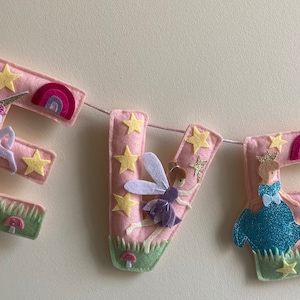 K&ouml;nnte beinhalten: Ein Filzbuchstaben-Banner, das EVE in Hellrosa buchstabiert, mit einem wei&szlig;en Einhorn auf dem E, einer Fee auf dem V und einer Prinzessin im blauen Kleid auf dem E. Jeder Buchstabe ist mit gelben Sternen, kleinen Regenb&ouml;gen, Gras und Pilzen verziert.