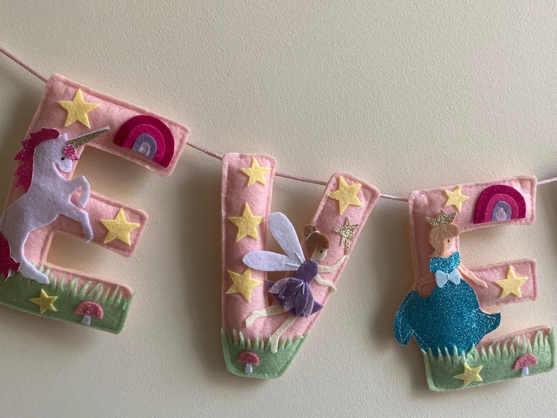 K&ouml;nnte beinhalten: Ein Filzbuchstaben-Banner, das EVE in Hellrosa buchstabiert, mit einem wei&szlig;en Einhorn auf dem E, einer Fee auf dem V und einer Prinzessin im blauen Kleid auf dem E. Jeder Buchstabe ist mit gelben Sternen, kleinen Regenb&ouml;gen, Gras und Pilzen verziert.