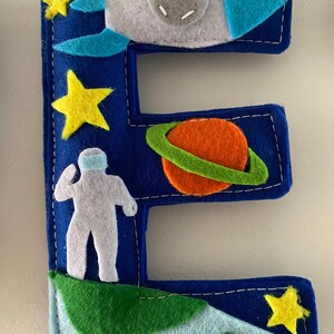 May include: A dark blue felt letter E decorated with space-themed appliqu&eacute;s including a grey rocket ship, a bright orange planet with a green ring, a white astronaut figure, and several yellow stars, all stitched with white thread onto the letter.