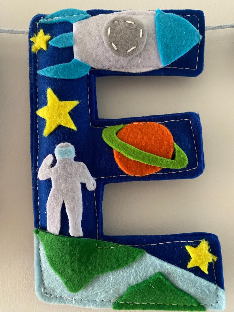 May include: A dark blue felt letter E decorated with space-themed appliqu&eacute;s including a grey rocket ship, a bright orange planet with a green ring, a white astronaut figure, and several yellow stars, all stitched with white thread onto the letter.