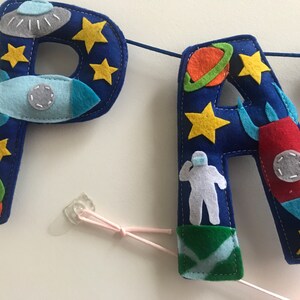 May include: A blue felt banner featuring the letters P and A decorated with space-themed appliqu&eacute;s including yellow stars, a gray UFO, a light blue rocket, a red rocket, a green planet with an orange ring, and a white astronaut figure on a green base.