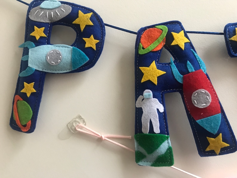 May include: A blue felt banner featuring the letters P and A decorated with space-themed appliqu&eacute;s including yellow stars, a gray UFO, a light blue rocket, a red rocket, a green planet with an orange ring, and a white astronaut figure on a green base.