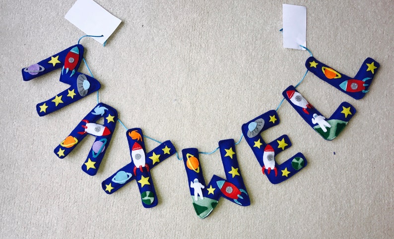 May include: A hanging banner spelling the name MAXWELL in dark blue felt letters. Each letter is decorated with space-themed appliqu&eacute;s including rockets, planets, stars, astronauts, and flying saucers, all connected by a thin light blue string.