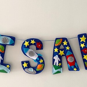 May include: A decorative banner spelling the name ISAK in dark blue felt letters. Each letter is adorned with space-themed appliqu&eacute;s including yellow stars, colorful planets, white astronauts, and small rockets, with two larger rockets on the ends.