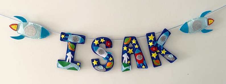 May include: A decorative banner spelling the name ISAK in dark blue felt letters. Each letter is adorned with space-themed appliqu&eacute;s including yellow stars, colorful planets, white astronauts, and small rockets, with two larger rockets on the ends.