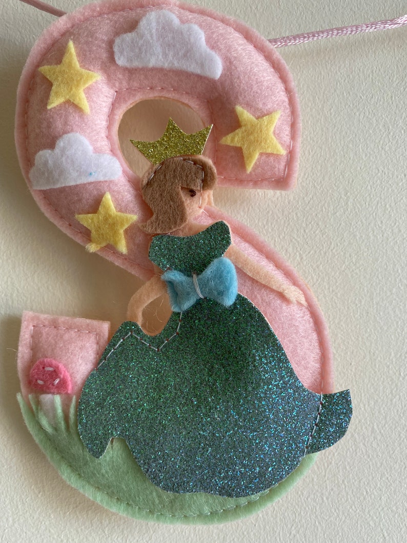 K&ouml;nnte beinhalten: Ein Buchstabe S aus Filz in Hellrosa, verziert mit gelben Sternen und wei&szlig;en Wolken. Eine Figur mit braunen Haaren, goldener Krone und einem gr&uuml;nen Glitzerkleid mit blauer Schleife steht vor dem Buchstaben. Ein kleiner rosa Pilz auf gr&uuml;nem Gras.