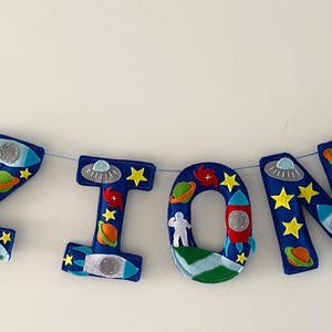 May include: A felt banner spelling the name ZION in large blue letters, decorated with space-themed appliqu&eacute;s including rockets, planets, stars, astronauts, and UFOs, strung on a thin cord with two additional rocket shapes at the ends.