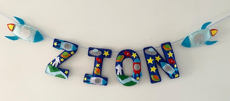 May include: A felt banner spelling the name ZION in large blue letters, decorated with space-themed appliqu&eacute;s including rockets, planets, stars, astronauts, and UFOs, strung on a thin cord with two additional rocket shapes at the ends.