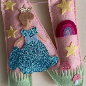 K&ouml;nnte beinhalten: Ein Buchstabe N aus rosa Filz, verziert mit einer glitzernden blauen Figur in einem Kleid mit goldener Krone, gelben Sternen, einem kleinen Regenbogen und zwei rosa Pilzen in hellgr&uuml;nem Filzgras. Das St&uuml;ck ist mit wei&szlig;em Garn vern&auml;ht.