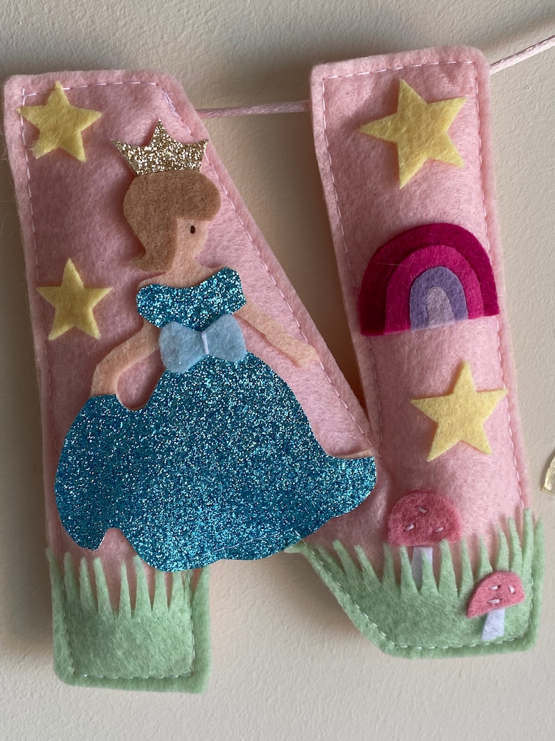 K&ouml;nnte beinhalten: Ein Buchstabe N aus rosa Filz, verziert mit einer glitzernden blauen Figur in einem Kleid mit goldener Krone, gelben Sternen, einem kleinen Regenbogen und zwei rosa Pilzen in hellgr&uuml;nem Filzgras. Das St&uuml;ck ist mit wei&szlig;em Garn vern&auml;ht.
