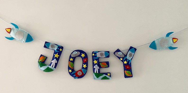 May include: A decorative banner spelling the name JOEY in dark blue felt letters. Each letter is adorned with colorful space-themed felt appliqu&eacute;s including rockets, planets, stars, and astronauts, with two additional rocket cutouts on the ends.