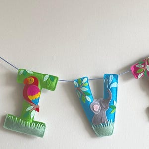 Könnte beinhalten: Ein hängendes Banner, das den Namen OLIVIA mit einzelnen Filzbuchstaben buchstabiert. Jeder Buchstabe zeigt Dschungeltier-Applikationen wie Nilpferd, Giraffe, Papagei, Elefant, Löwe und Affe, akzentuiert mit grünen Blatt- und Grasmotiven.