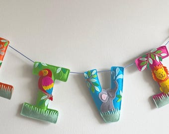 May include: A hanging banner spelling the name OLIVIA with individual felt letters. Each letter features jungle animal appliques including a hippo, giraffe, parrot, elephant, lion, and monkey, accented with green leaves and grass motifs.