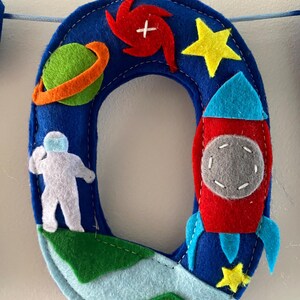 May include: A blue felt letter O decoration featuring space-themed appliqu&eacute;s including a green planet with an orange ring, a red spiral galaxy, a white astronaut, a red and blue rocket ship, and two yellow stars, all stitched onto a blue base.