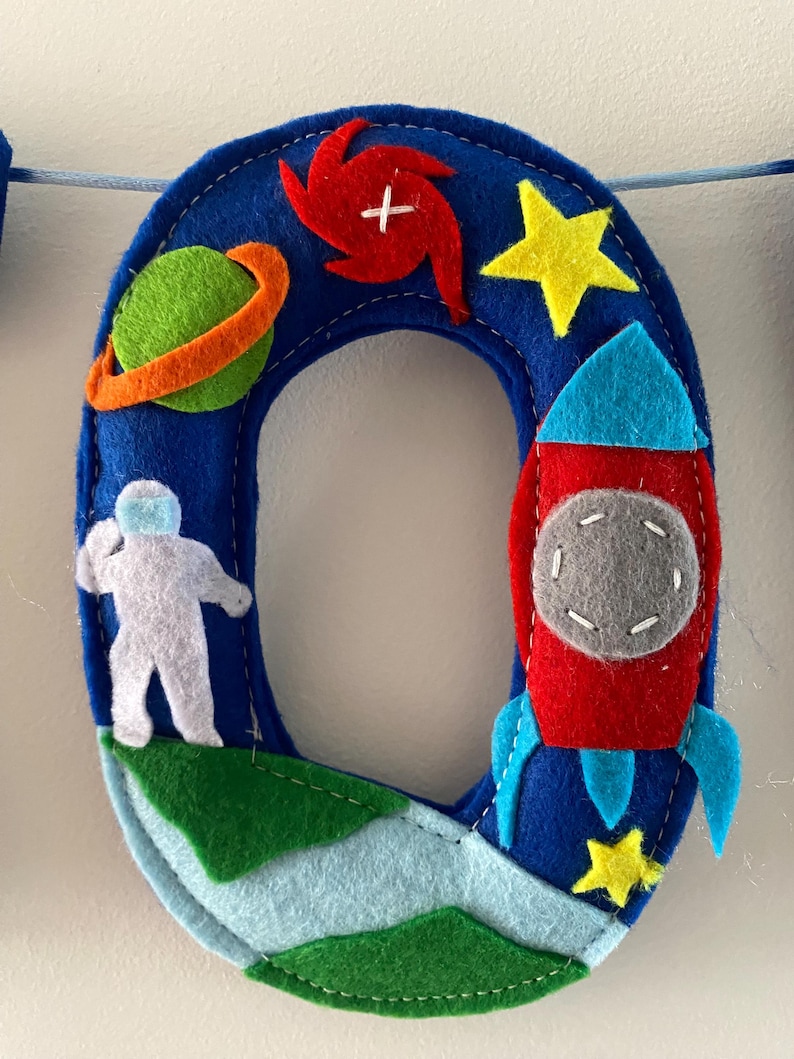 May include: A blue felt letter O decoration featuring space-themed appliqu&eacute;s including a green planet with an orange ring, a red spiral galaxy, a white astronaut, a red and blue rocket ship, and two yellow stars, all stitched onto a blue base.
