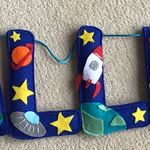 May include: A felt banner spelling the name CALLUM in deep blue letters, decorated with space-themed appliqu&eacute;s including yellow stars, orange and blue planets, white rockets, gray flying saucers, green earth-like globes, and white astronauts.