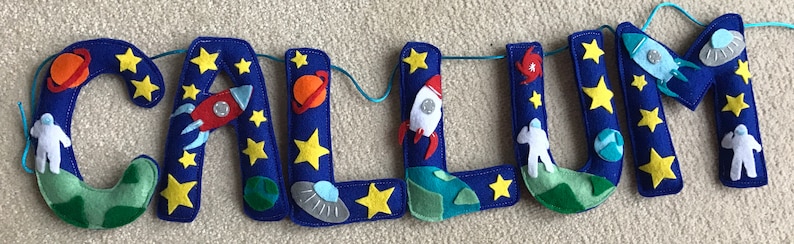 May include: A felt banner spelling the name CALLUM in deep blue letters, decorated with space-themed appliqu&eacute;s including yellow stars, orange and blue planets, white rockets, gray flying saucers, green earth-like globes, and white astronauts.