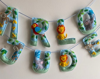 May include: A felt letter banner with the name "ARLO JUDE" spelled out in blue letters. Each letter is decorated with a different animal, including a monkey, parrot, lion, giraffe, elephant, and a monkey.