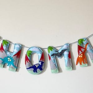 Könnte beinhalten: Ein Filzbanner mit dem Namen "THOMAS" in blauen Buchstaben. Jeder Buchstabe hat einen Dinosaurier und eine Bergszene. Die Dinosaurier sind orange, gelb, blau und grün.