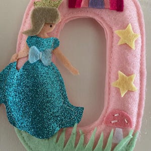 K&ouml;nnte beinhalten: Eine Filzdekoration in Form der Zahl Null in Hellrosa, verziert mit einer glitzernden blauen Prinzessinnenfigur mit goldener Krone, einem kleinen Regenbogen, gelben Sternen, einem rosa Pilz und hellgr&uuml;nen Grasakzenten auf der Vorderseite.
