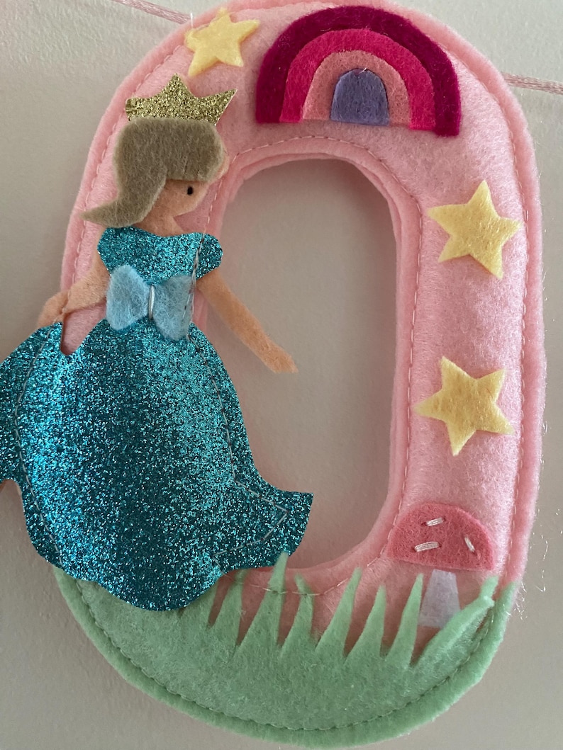 K&ouml;nnte beinhalten: Eine Filzdekoration in Form der Zahl Null in Hellrosa, verziert mit einer glitzernden blauen Prinzessinnenfigur mit goldener Krone, einem kleinen Regenbogen, gelben Sternen, einem rosa Pilz und hellgr&uuml;nen Grasakzenten auf der Vorderseite.