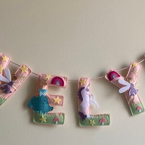 K&ouml;nnte beinhalten: Ein Filzbuchstaben-Banner, das EVELYN in Hellrosa buchstabiert, mit dekorativen Applikationen aus Einh&ouml;rnern, Feen, Regenb&ouml;gen, gelben Sternen, gr&uuml;nen Grasfl&auml;chen und kleinen roten Pilzen, die an jedem einzelnen Buchstaben befestigt sind.