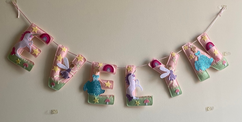 K&ouml;nnte beinhalten: Ein Filzbuchstaben-Banner, das EVELYN in Hellrosa buchstabiert, mit dekorativen Applikationen aus Einh&ouml;rnern, Feen, Regenb&ouml;gen, gelben Sternen, gr&uuml;nen Grasfl&auml;chen und kleinen roten Pilzen, die an jedem einzelnen Buchstaben befestigt sind.