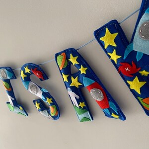 May include: A felt banner spelling the name SAM with blue letters decorated with space-themed appliqu&eacute;s including yellow stars, red and orange planets, white astronauts, and light blue rockets, all strung together on a thin light blue cord.
