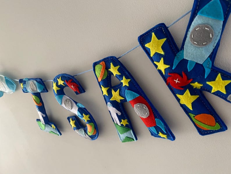 May include: A felt banner spelling the name SAM with blue letters decorated with space-themed appliqu&eacute;s including yellow stars, red and orange planets, white astronauts, and light blue rockets, all strung together on a thin light blue cord.