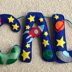 May include: A felt letter banner spelling the name CAL with a space theme. The blue letters are decorated with yellow stars, an orange planet with a ring, a red rocket ship, a white astronaut, a green planet, and a gray flying saucer.
