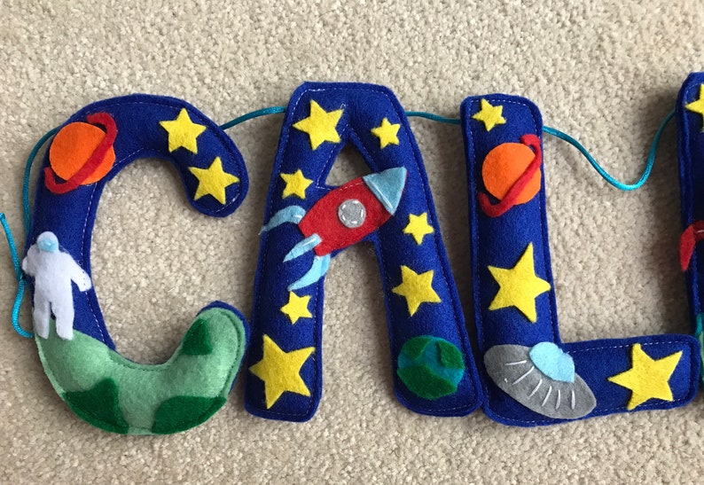 May include: A felt letter banner spelling the name CAL with a space theme. The blue letters are decorated with yellow stars, an orange planet with a ring, a red rocket ship, a white astronaut, a green planet, and a gray flying saucer.