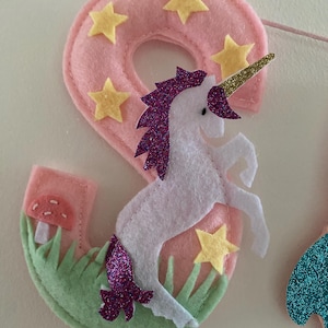 K&ouml;nnte beinhalten: Eine Filzdekoration in Form der Zahl Neun in Hellrosa mit gelben Sternausschnitten, einem wei&szlig;en Einhorn mit violetter Glitzerm&auml;hne und -schweif, einem goldenen Glitzerhorn, gr&uuml;nem Filzgras und einem kleinen roten Pilz mit wei&szlig;en Punkten.
