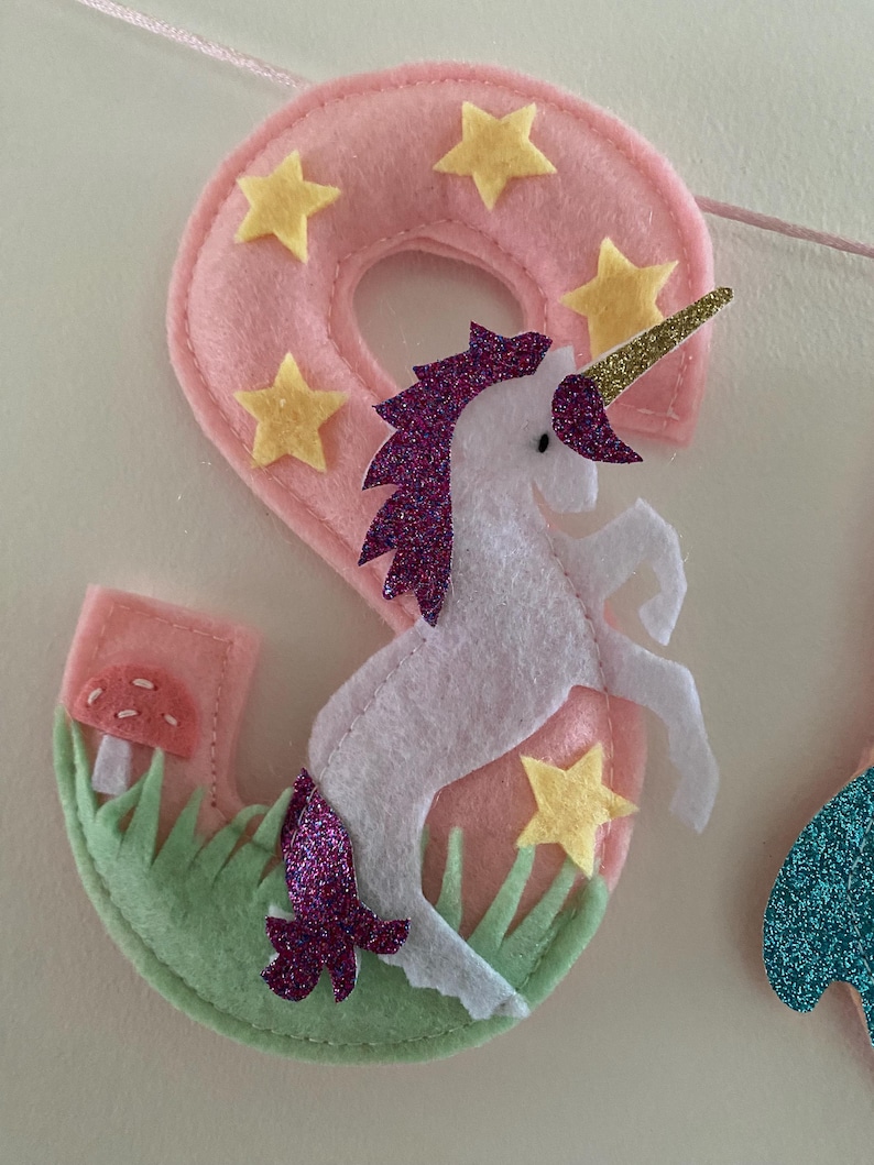 K&ouml;nnte beinhalten: Eine Filzdekoration in Form der Zahl Neun in Hellrosa mit gelben Sternausschnitten, einem wei&szlig;en Einhorn mit violetter Glitzerm&auml;hne und -schweif, einem goldenen Glitzerhorn, gr&uuml;nem Filzgras und einem kleinen roten Pilz mit wei&szlig;en Punkten.