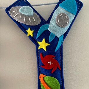 May include: A dark blue felt letter Y decoration featuring space-themed appliqu&eacute;s including a grey UFO, a light blue rocket ship, two yellow stars, a red spiral galaxy, and a green planet with an orange ring, all stitched with white thread.