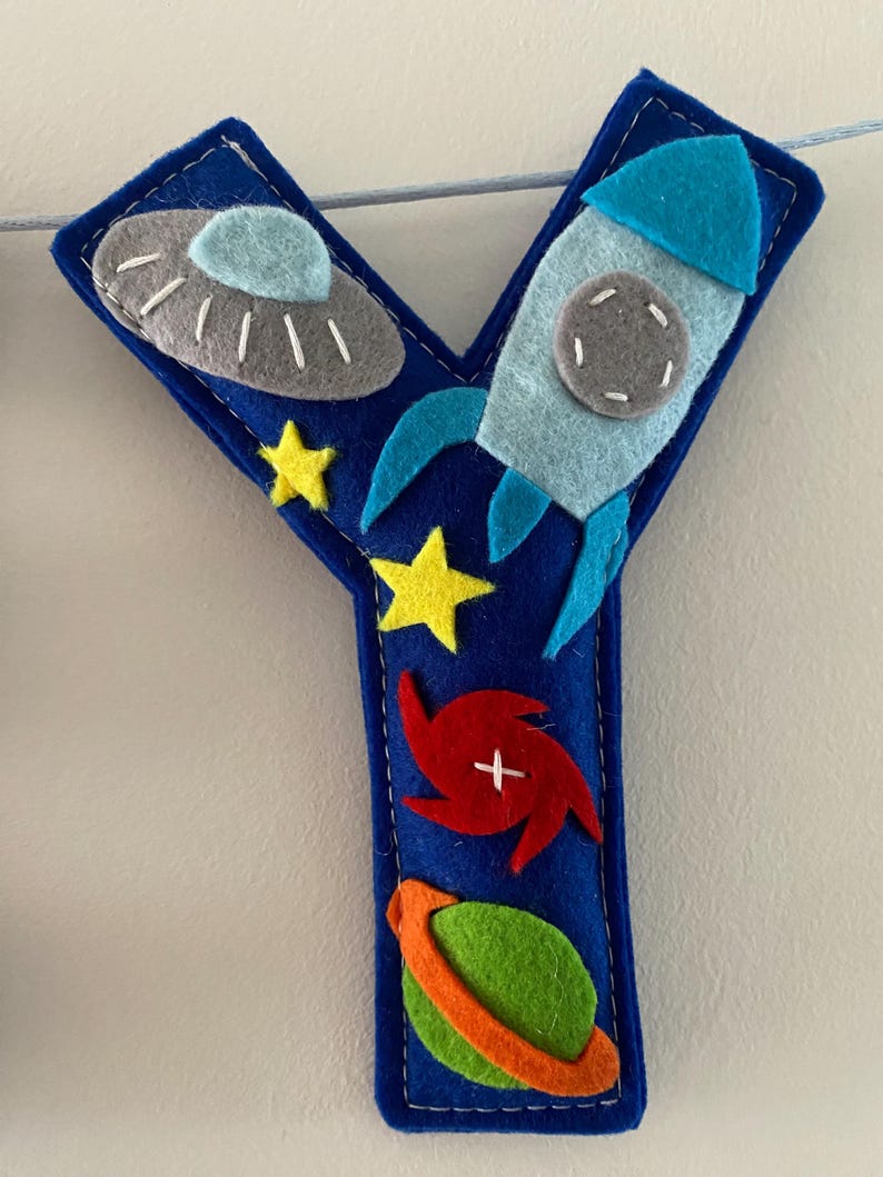 May include: A dark blue felt letter Y decoration featuring space-themed appliqu&eacute;s including a grey UFO, a light blue rocket ship, two yellow stars, a red spiral galaxy, and a green planet with an orange ring, all stitched with white thread.