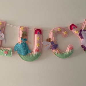 K&ouml;nnte beinhalten: Ein Filzbuchstaben-Banner, das LUCY in Hellrosa buchstabiert, mit dekorativen Applikationen wie wei&szlig;en Einh&ouml;rnern mit lila M&auml;hnen, einer Prinzessin im blauen Kleid, einer Fee mit Fl&uuml;geln, gelben Sternen, Regenb&ouml;gen, gr&uuml;nem Gras und kleinen roten Pilzen.