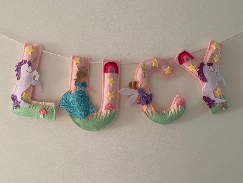 K&ouml;nnte beinhalten: Ein Filzbuchstaben-Banner, das LUCY in Hellrosa buchstabiert, mit dekorativen Applikationen wie wei&szlig;en Einh&ouml;rnern mit lila M&auml;hnen, einer Prinzessin im blauen Kleid, einer Fee mit Fl&uuml;geln, gelben Sternen, Regenb&ouml;gen, gr&uuml;nem Gras und kleinen roten Pilzen.