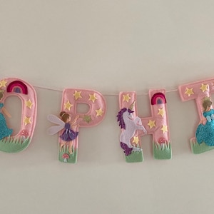 K&ouml;nnte beinhalten: Ein dekoratives Banner, das den Namen SOPHTA in rosa Filzbuchstaben buchstabiert. Jeder Buchstabe ist mit gelben Sternen, gr&uuml;nem Gras und Figuren wie Einh&ouml;rnern, Feen und blau glitzernden Kleidern verziert, die an einer d&uuml;nnen wei&szlig;en Schnur h&auml;ngen.