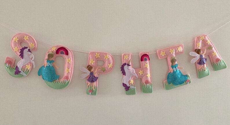 K&ouml;nnte beinhalten: Ein dekoratives Banner, das den Namen SOPHTA in rosa Filzbuchstaben buchstabiert. Jeder Buchstabe ist mit gelben Sternen, gr&uuml;nem Gras und Figuren wie Einh&ouml;rnern, Feen und blau glitzernden Kleidern verziert, die an einer d&uuml;nnen wei&szlig;en Schnur h&auml;ngen.