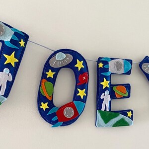 May include: A felt letter banner spelling the name JOEY in dark blue, featuring space-themed appliqu&eacute;s including rockets, planets, astronauts, stars, and flying saucers. The letters are strung together on a thin white cord with a small rocket accent.
