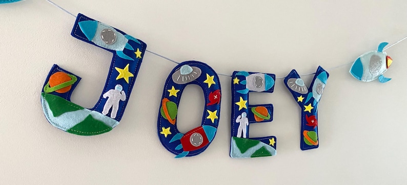 May include: A felt letter banner spelling the name JOEY in dark blue, featuring space-themed appliqu&eacute;s including rockets, planets, astronauts, stars, and flying saucers. The letters are strung together on a thin white cord with a small rocket accent.